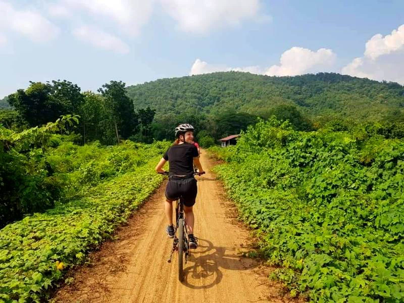 Een vrouw fietst over een zandpad omringd door groene struiken. Op de achtergrond zijn beboste heuvels en een blauwe lucht met wat wolken te zien.