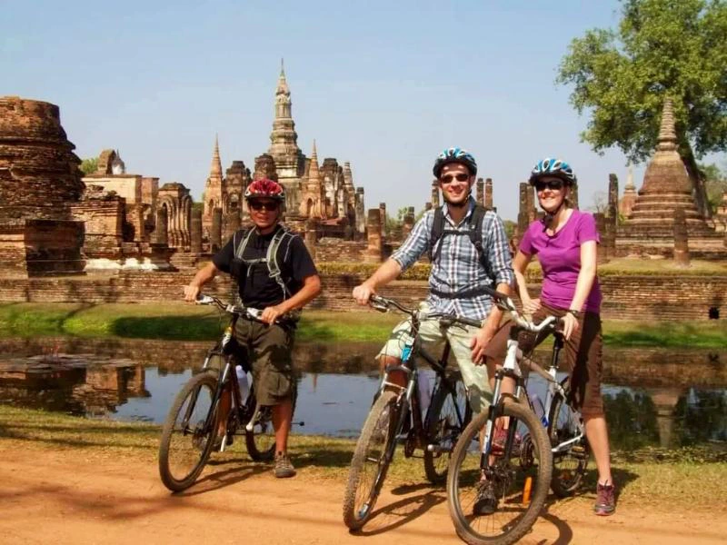 Drie mensen met fietsen staan op een zandpad bij een waterpartij. Op de achtergrond zie je oude tempelruïnes met torens en stoepa’s van baksteen en steen, en een boom links in beeld.