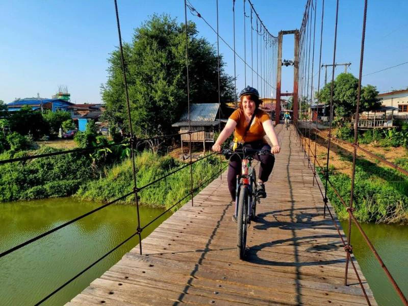 Een vrouw fietst over een smalle houten hangbrug met touwen aan weerszijden. Onder de brug stroomt een rivier en op de achtergrond zie je huizen, bomen en elektriciteitspalen.
