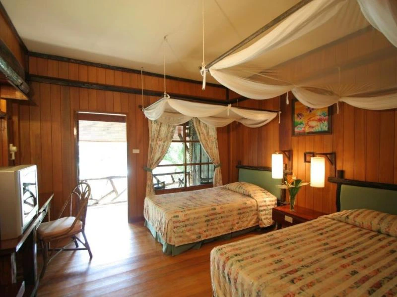 Hotelkamer met twee eenpersoonsbedden, houten muren en een wit plafond. Boven elk bed hangt een klamboe. In de kamer staat een klein bureautje met een stoel en een kluis.