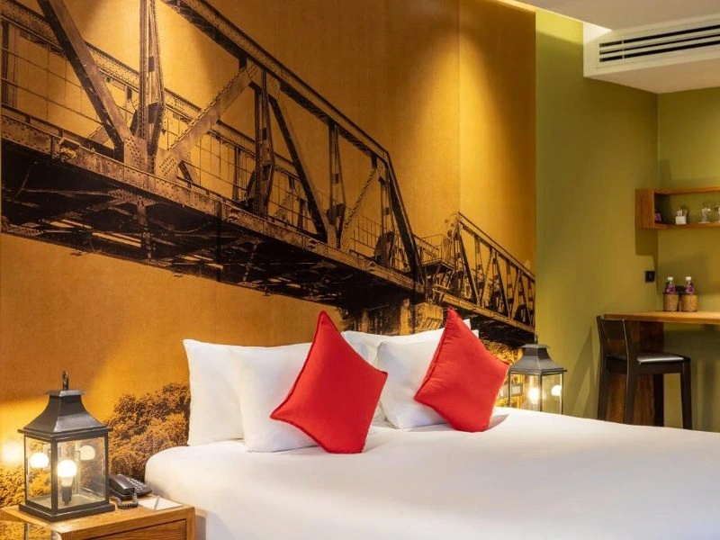 Stijlvolle hotelkamer met groot bed, rode kussens en sfeerverlichting, met een grote muurfoto van de Brug over de River Kwai als eyecatcher.