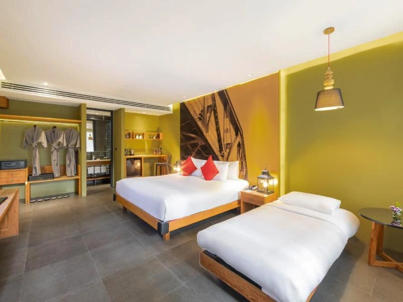 Hotelkamer met een tweepersoonsbed met ronde sierkussens, een eenpersoonsbed, groene wanden, een grote foto aan de muur, een minibar en een open kast met hangende badjassen.
