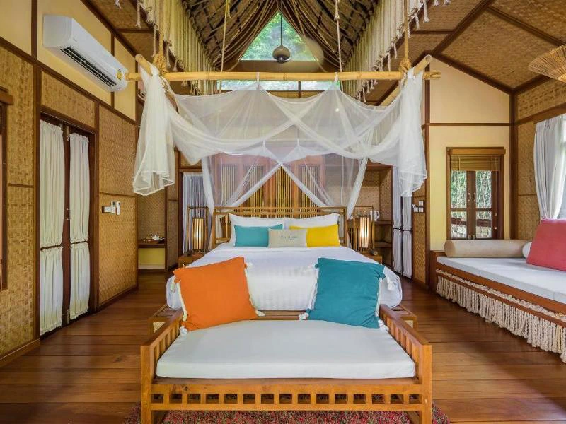 Binnenkant van een bamboe bungalow met een tweepersoonsbed met klamboe, een bankje aan het voeteneind, een loungehoek bij het raam en uitzicht op de jungle