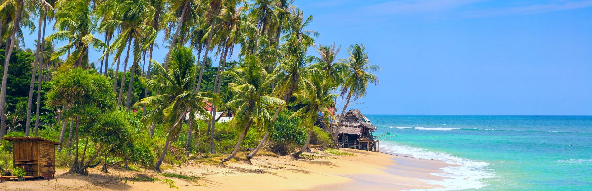 een strand op Koh Lanta, met palmbomen die wuiven in de wind, golven die in de branding stukslaan op het strand en een helderblauwe zee daarachter
