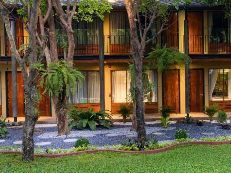 Twee verdiepingen tellend hotelgebouw met houten deuren en grote ramen met gordijnen, omringd door een verzorgde tuin met bomen, sierplanten en een pad van stapstenen op een ondergrond van grijze kiezelstenen.