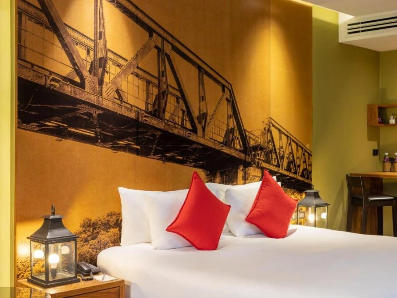 Tweepersoonsbed met witte lakens en rode kussens, grote muurfoto van een stalen brug op een okergele achtergrond.