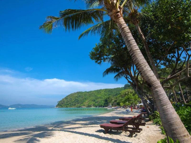 Zandstrand met ligbedden onder palmbomen, uitkijkend over een turquoise zee en groene heuvels.