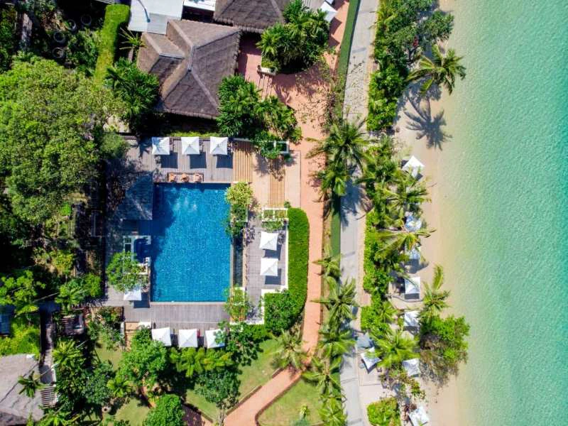 Luchtfoto van zwembad met ligbedden en parasols, omgeven door palmbomen en gelegen naast een helder turquoise strand.