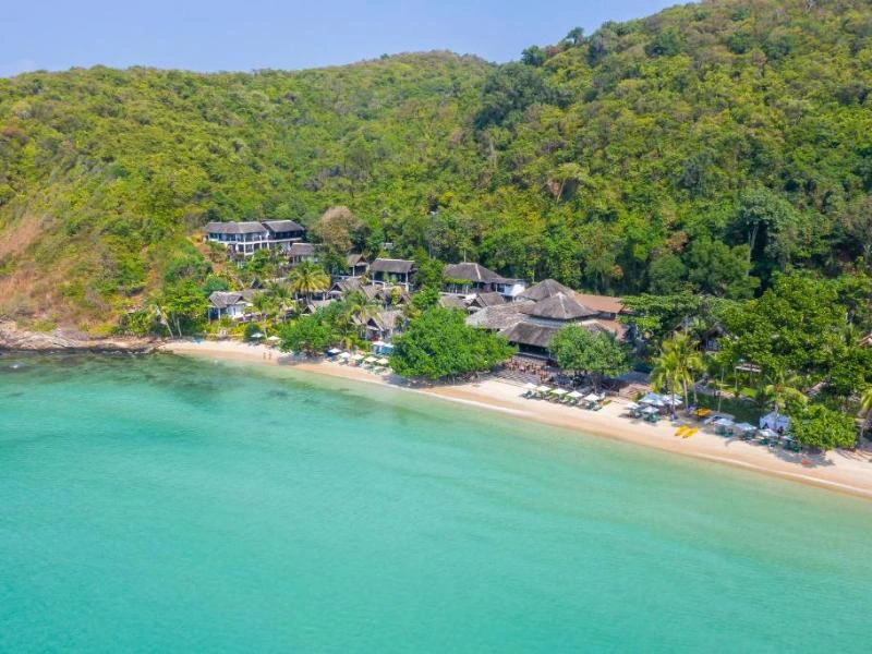 Azuurblauw water en een rustig strand, omringd door tropisch groen met een kleinschalig resort verscholen tussen de bomen.