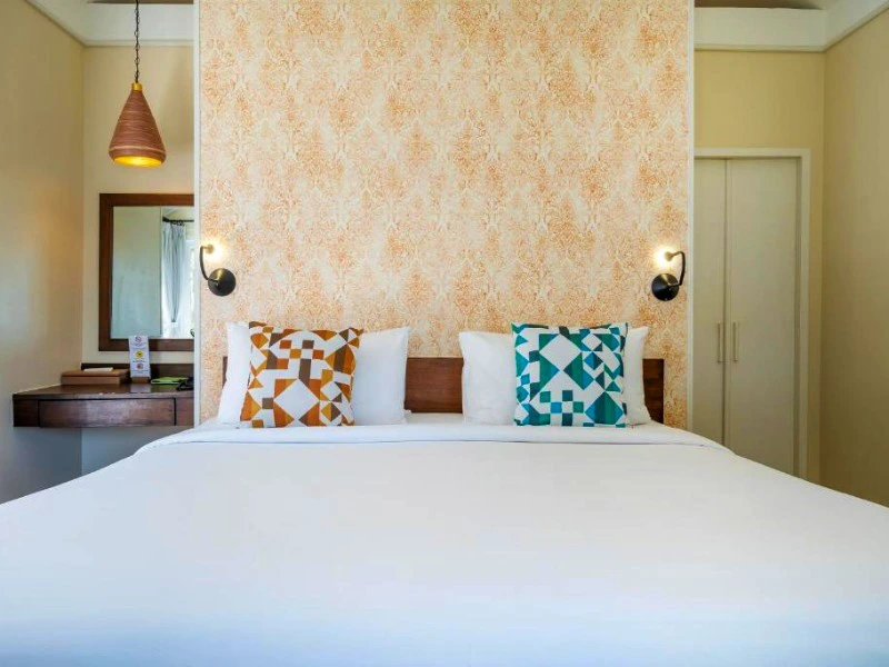 Comfortabel tweepersoonsbed met decoratieve kussens en warme verlichting, tegen een zacht patroonbehang.