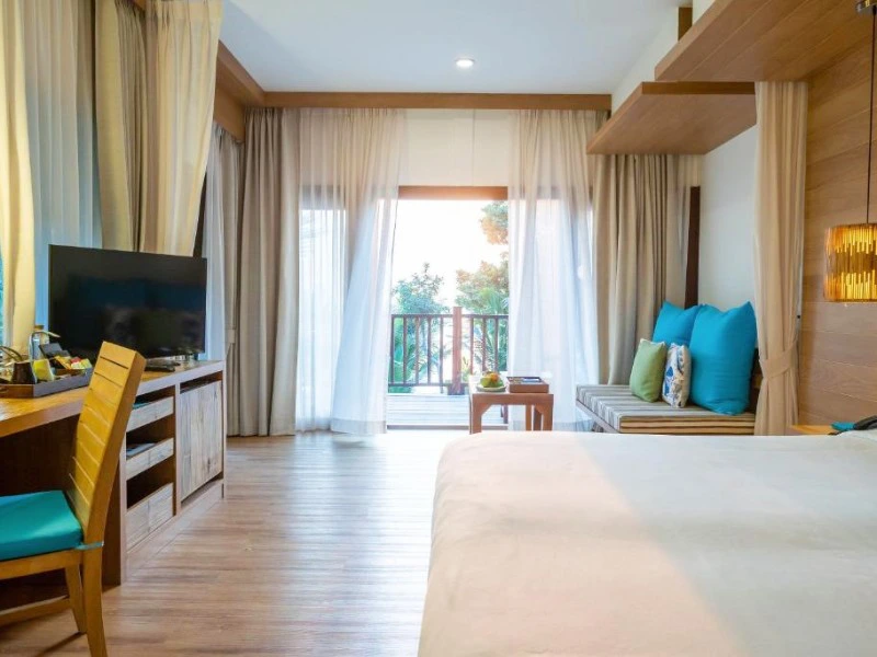 Lichte hotelkamer met houten accenten, een ruim bed, zithoek met kleurrijke kussens en een balkon met uitzicht op tropisch groen.