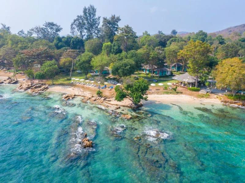 Rotsachtige kustlijn met kristalhelder turquoise water, een rustig strand en verspreide ligstoelen onder de schaduw van groene bomen.