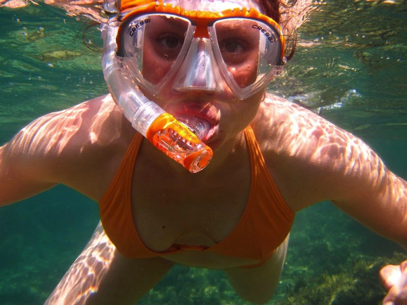 Vrouw met oranje snorkel en duikbril onder water, kijkt recht in de camera.