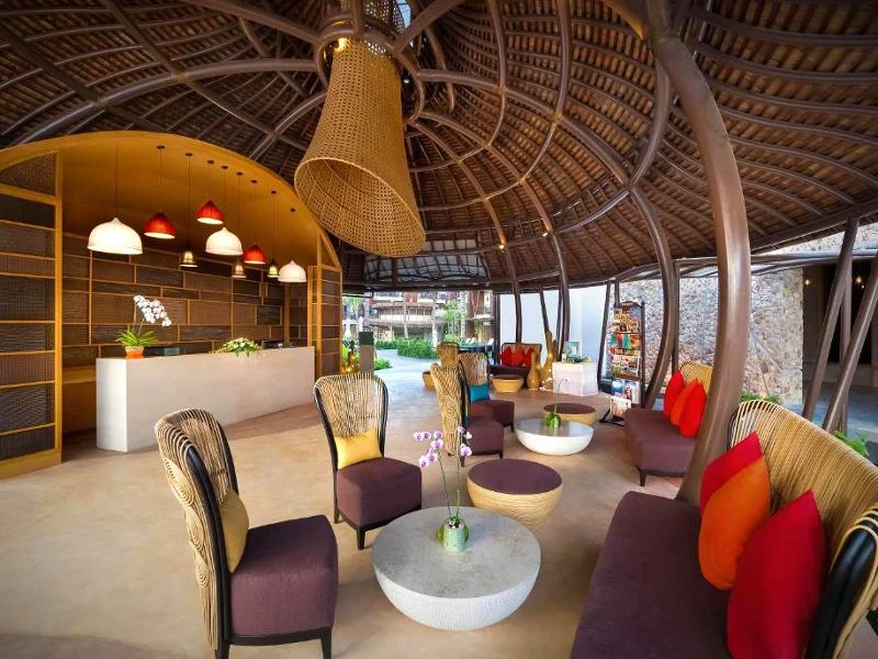 Open lobby met een rond gevormd houten dak en grote hanglamp. Er staan rotan stoelen en banken met felgekleurde kussens rondom ronde tafeltjes. Op de achtergrond is een receptiebalie te zien met rode en witte lampen erboven