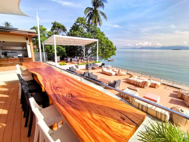 Terras met zitzakken en een open bar onder een overkapping, met daarachter begroeiing. Uitzicht op zee met een klein eilandje in de verte en een paar wolken in de lucht