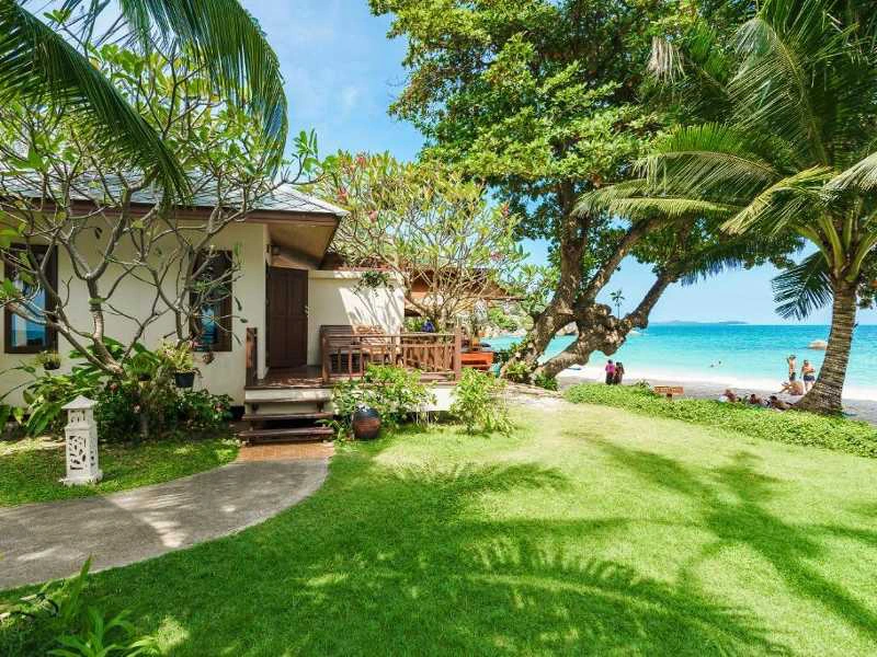 Bungalow aan zee met groene tuin, palmbomen en uitzicht op strand en helderblauw water.
