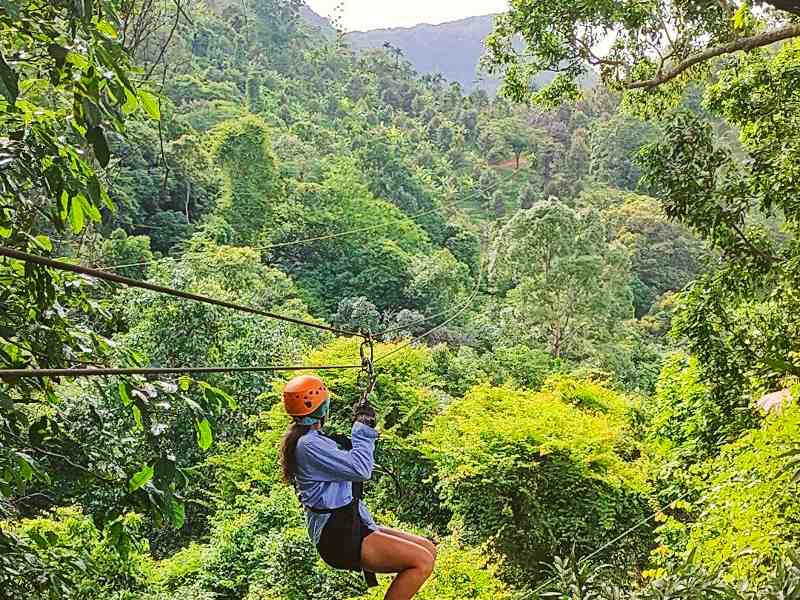 Vrouw met helm suist op een zipline door een dichtbegroeid, groen berglandschap.