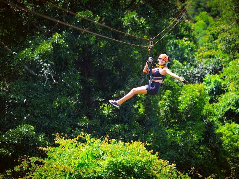 Vrouw in sportkleding zweeft lachend aan een zipline boven een groene jungle.