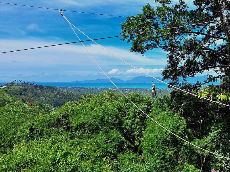 Persoon op een zipline boven het groene regenwoud met uitzicht op zee en bergen.