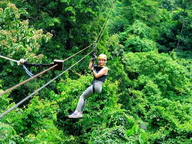 Vrouwelijke reiziger met oranje helm hangt in een tuigje aan een ziplijn boven een groene jungle.