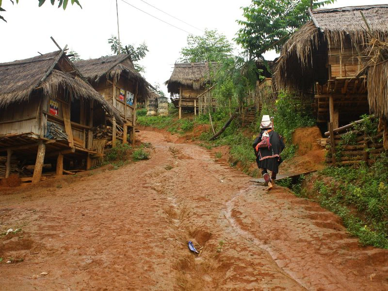 Een vrouw in traditionele kleding loopt een modderig pad op, langs houten huizen op palen met rieten daken. De lucht is bewolkt.
