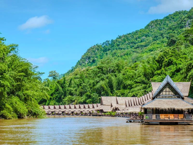 Rijtje houten junglehuisjes op een vlot aan de rivier, omringd door groene heuvels en tropisch regenwoud, helder blauwe lucht.