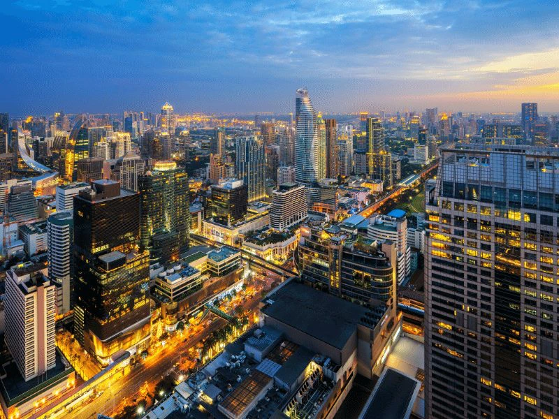 Uitzicht over de skyline van Bangkok tijdens de schemering, met hoge gebouwen, verlichte straten en een kleurrijke avondlucht. De stad bruist van leven en moderne architectuur.