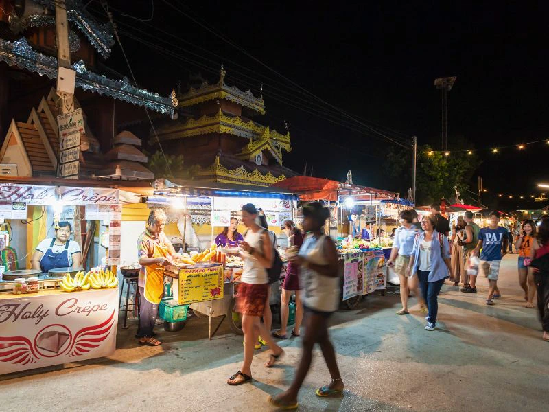 De foto toont een avondmarkt met verlichte eetkraampjes, waar onder andere fruit en crêpes worden verkocht. Op de achtergrond staat een tempelachtig gebouw met gouden details en rood dak. Er lopen mensen op slippers, sandalen of blote voeten. Boven hangen lampjes en stroomkabels.