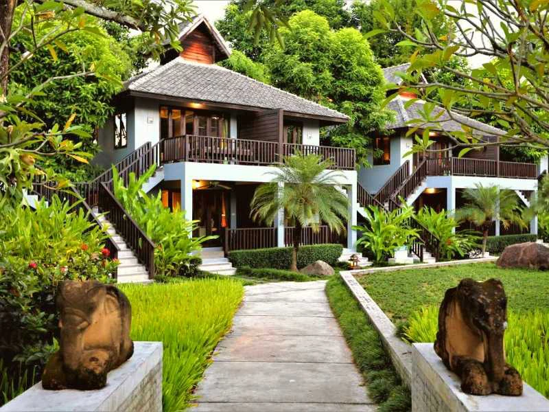 Twee witte guesthouses tussen dichte groene begroeiing, met trappen naar de eerste verdieping. Balustrades en kozijnen van donker hout. Tussen de huizen loopt een pad met bij de ingang aan beide zijden beelden.