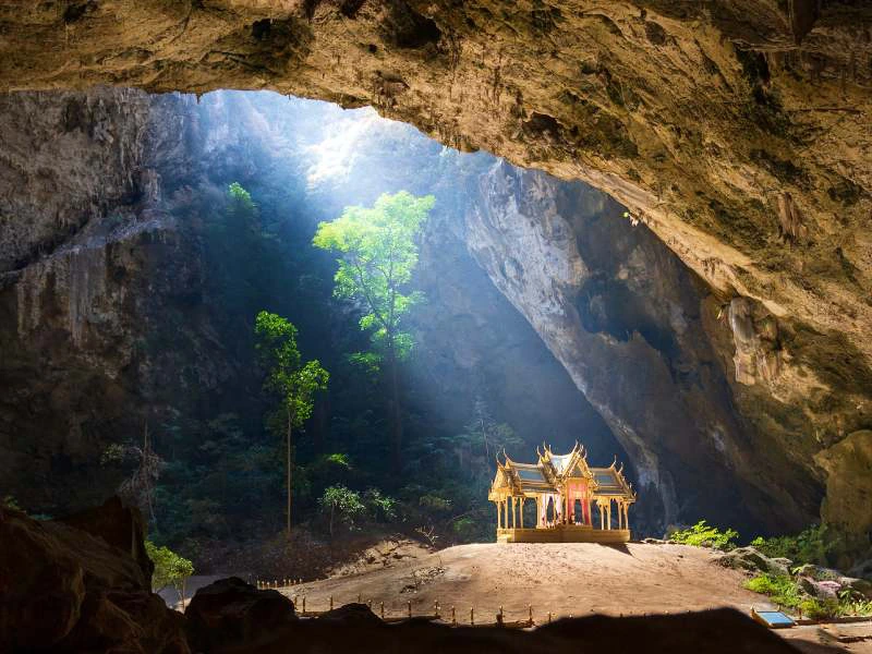 Traditioneel Thais paviljoen verlicht door een zonnestraal in een grote grot, omringd door rotswanden en hoge bomen.