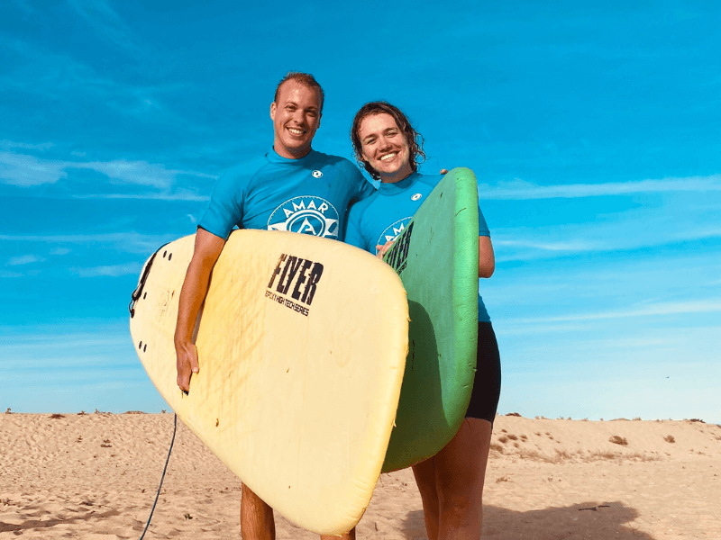 Twee reizigers staan lachend op het strand, gezicht naar de camera, met elk een surfplank en een blauw shirt aan. Lucht is helder.