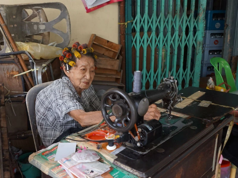 Een oudere Thaise vrouw zit buiten achter een klassieke naaimachine, geconcentreerd aan het werk. Ze draagt krulspelden in haar haar en zit in een eenvoudige, rommelige werkruimte met een groene schuifdeur op de achtergrond.