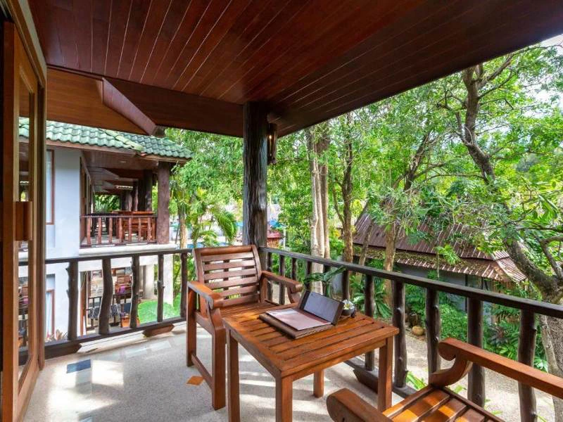 Overdekt balkon met houten stoelen en tafel, waarop een laptop en een map liggen, met uitzicht op bomen en omliggende huisjes in een tropische setting.