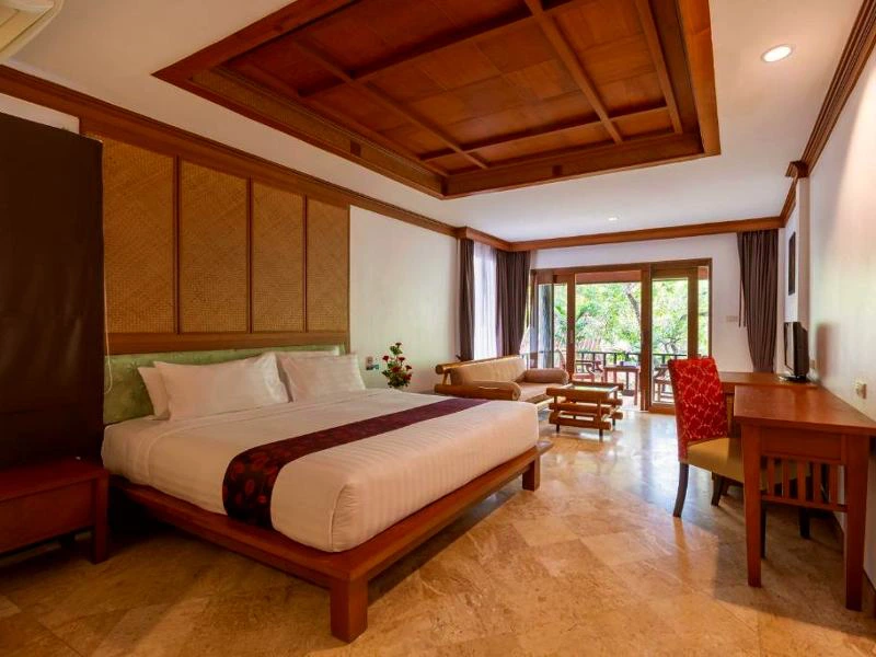 Ruime hotelkamer met houten plafond, groot tweepersoonsbed, zithoek met bank, bureau met stoelen en openslaande deuren naar een balkon met uitzicht op groen.