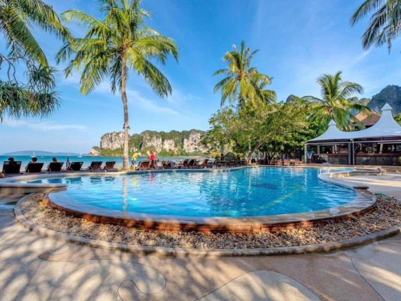 Rond zwembad met ligstoelen onder palmbomen, uitzicht op zee en kalkstenen kliffen op de achtergrond, naast een openluchtrestaurant met witte overkapping.