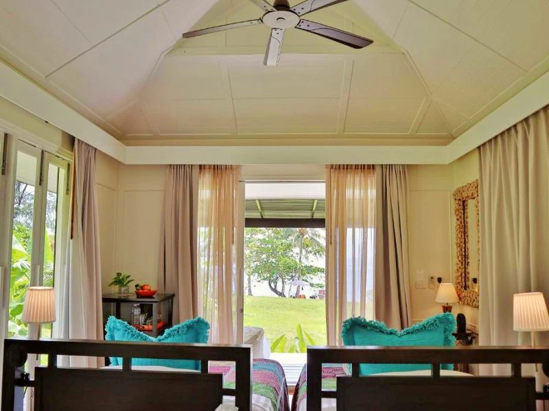 Hotelkamer met openstaande ramen en deuren met lichte gordijnen, twee losse bedden tegen elkaar met gekleurde dekbedovertrekken. Vanuit de kamer is uitzicht op de zee.