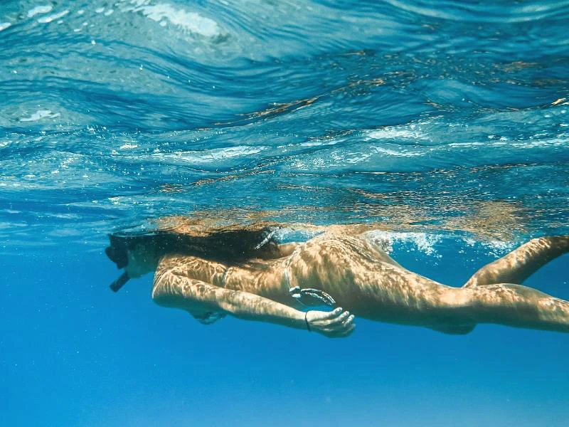 Onderwaterbeeld van een vrouw die snorkelend door helderblauw tropisch water zweeft, met zonlicht dat speels op haar huid en de wateroppervlakte danst.