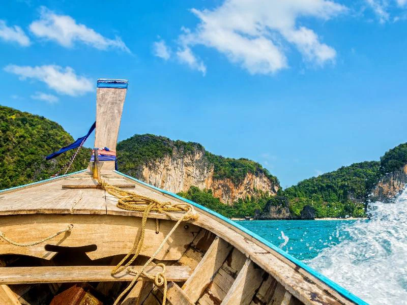 Voorsteven van een houten longtailboot op helderblauw water, met groene rotskliffen op de achtergrond en een zonnige lucht vol stapelwolken.