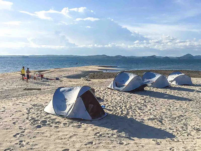 Vier uitklaptenten op een strand, met twee mensen die lopen. Zee en bergen zijn zichtbaar op de achtergrond