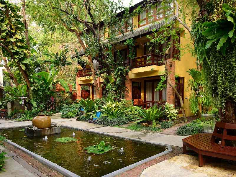 Tropische tuin met vijver en fontein bij een geel hotelgebouw met houten balkons.