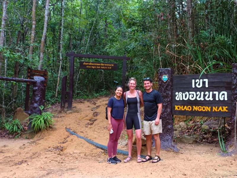 Drie wandelaars poseren lachend bij het startpunt van de Khao Ngon Nak trail in Thailand, met houten borden en jungle op de achtergrond.