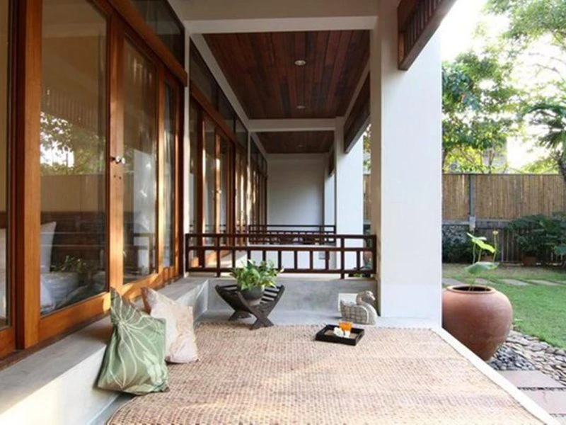 Een veranda met houten plafond en grote glazen schuifdeuren, ingericht met een rieten kleed, sierkussens en een lage tafel met drankjes. De veranda kijkt uit op een groene tuin met een grote aardewerken pot en bamboe schutting.