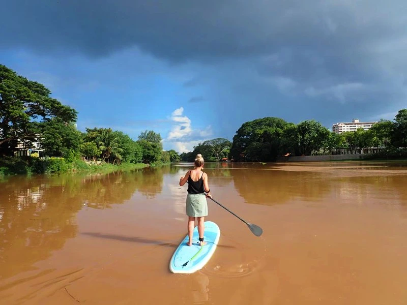 Reiziger staat op een blauw suppboard met een peddel in haar handen. De rivier is bruin van kleur, omringd door veel groen en een licht bewolkte lucht.