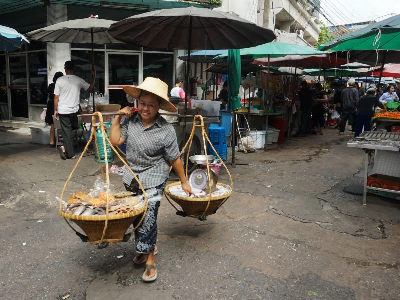 Een vrouw loopt op een marktstraat met twee gevlochten manden aan een draagstok over haar schouder. In de manden liggen etenswaren. Ze draagt een hoed en slippers. Op de achtergrond staan marktkramen met parasols en zijn andere mensen te zien.