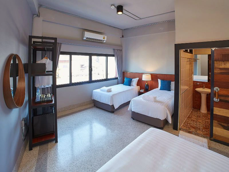 Een lichte hotelkamer met drie bedden: twee eenpersoonsbedden naast elkaar en één apart bed op de voorgrond. Er is een raam met gordijnen, een airco, een open badkamer met douche en toilet, en een kast met kleding en een waterkoker.