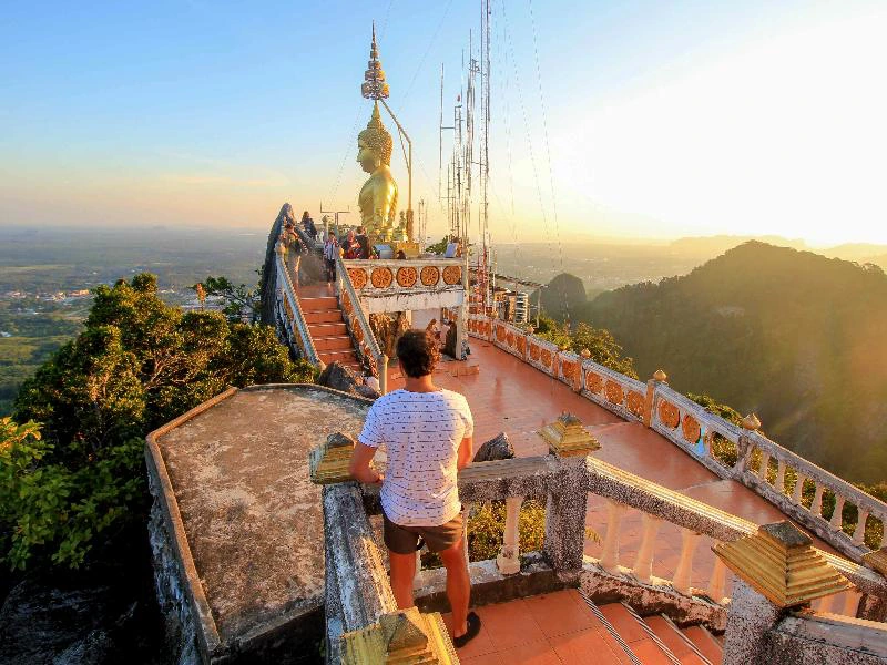 Man op uitkijkpunt bij een tempel met groot Boeddhabeeld op een bergtop, omringd door groene heuvels in de avondzon.