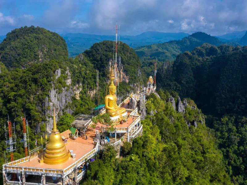 Gouden Boeddhabeeld en tempelcomplex op een bergtop, omringd door groene jungle en kalksteenrotsen onder een deels bewolkte blauwe lucht