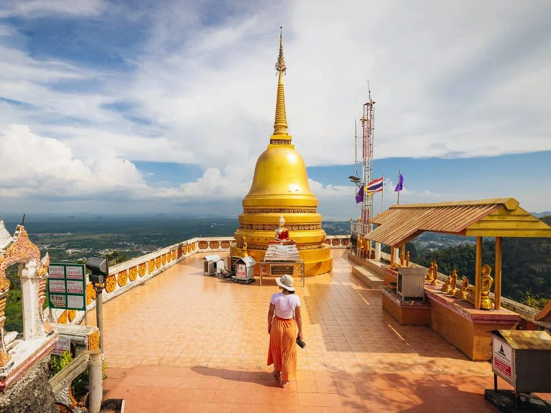 Uitzicht over een diepe vallei vanaf een betegeld platform van een tempelcomplex, met een reiziger op de voorgrond, licht bewolkte lucht op de achtergrond en rijk gedecoreerde tempelversieringen met gouden details.