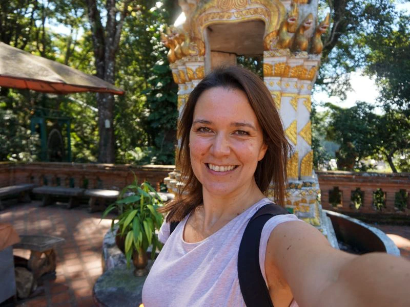 Vrouw maakt een selfie op een zonnige dag voor een rijk versierde tempelstructuur met goud en wit, omringd door bomen, planten en stenen bankjes.