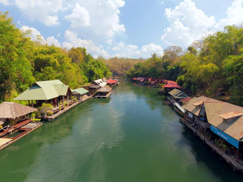 Houten huisjes met rieten of metalen daken drijven aan beide kanten van een brede rivier, omringd door groene bomen en heuvelachtige jungle.